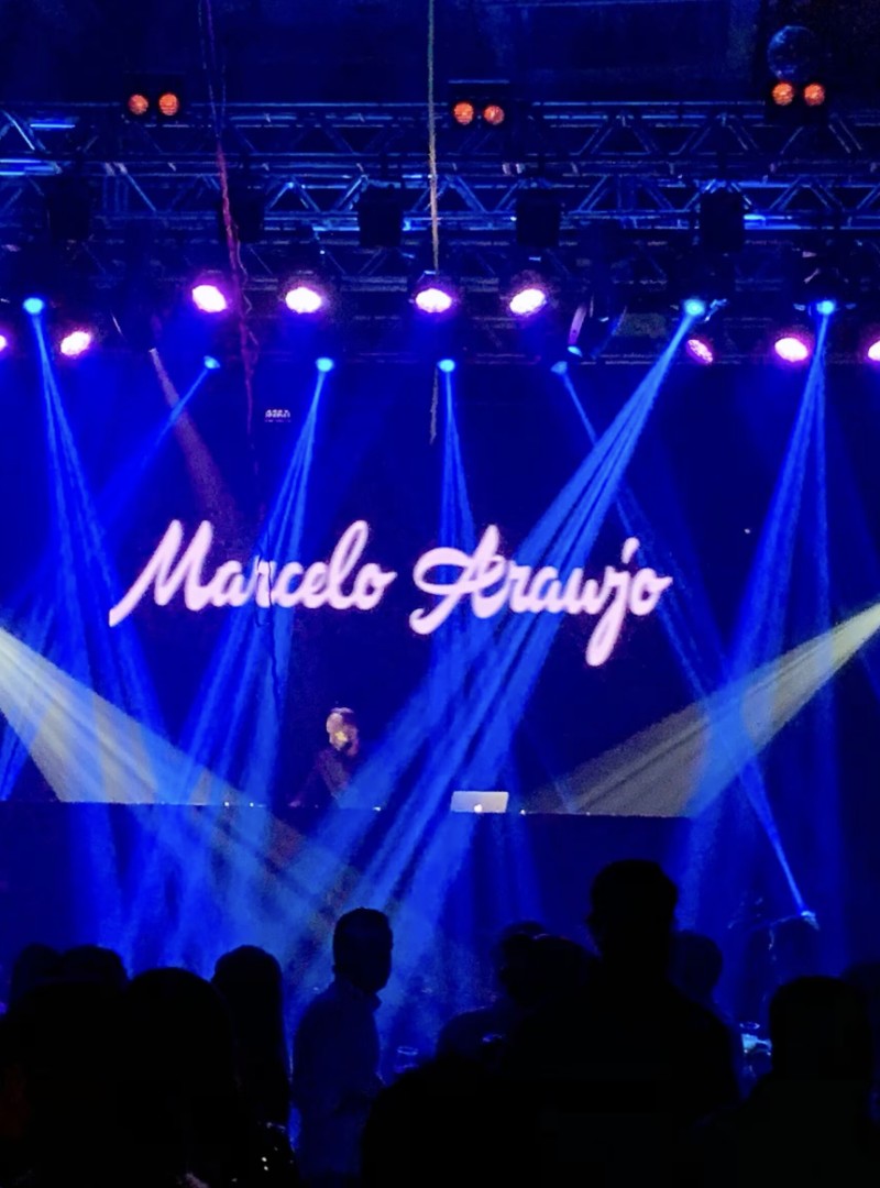 Marcelo Araujo tocando ao vivo em palco com seu nome iluminado em LED, público em frente
